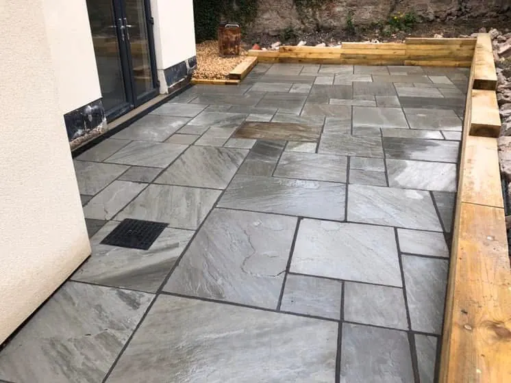 Grey stone slab patio with timber sleeper edging beside a rendered house. One of our jobs.